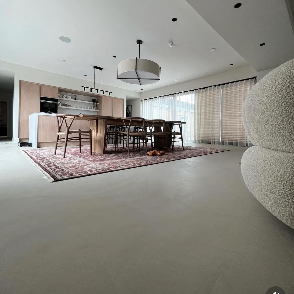 Open-concept kitchen and dining room with a long wooden table and chairs on a patterned rug, large windows with blinds, and a textured rounded lounge seat in the foreground.